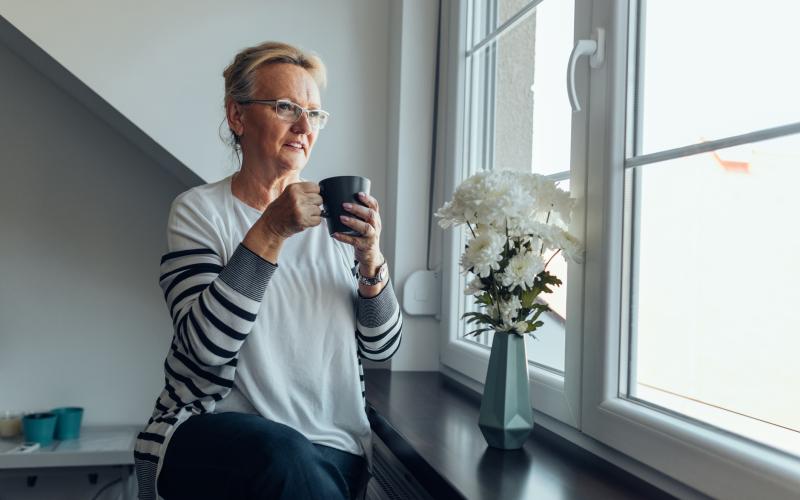 Senior vrouw zit bij het raam en heeft een mok in haar hand Senior vrouw zit bij het raam en heeft een mok in haar hand