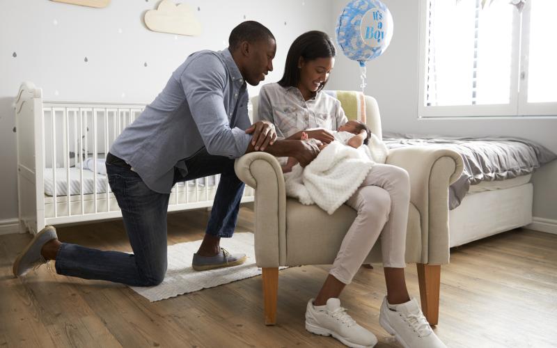 Vrouw zit met baby in haar armen op een stoel. Haar man zit geknield achter haar en kijkt glimlachend naar de baby. Vrouw zit met baby in haar armen op een stoel. Haar man zit geknield achter haar en kijkt glimlachend naar de baby.