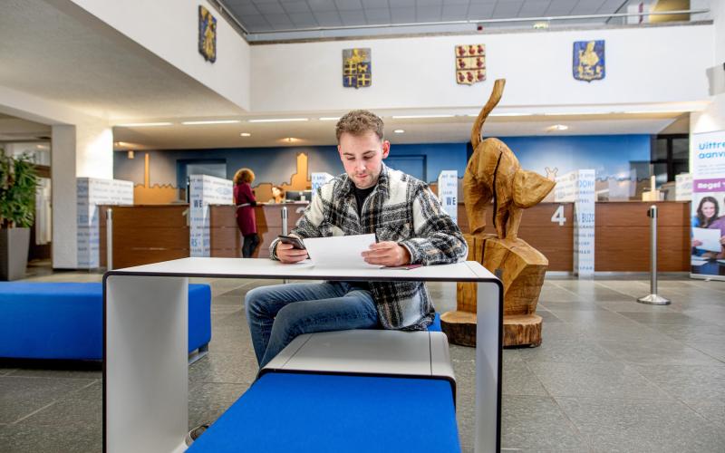 Een jongeman zit aan een tafel bij een gemeenteloket en vult een formulier in. Een jongeman zit aan een tafel bij een gemeenteloket en vult een formulier in.